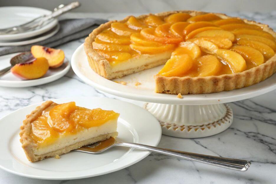 Classic peach tart with golden crust and glossy peach slices on white stand, served with a slice on a plate and silver fork.