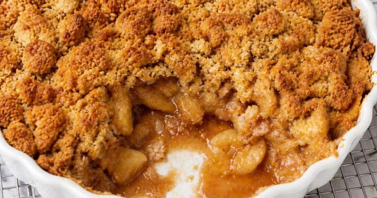 Close-up of a golden-brown apple caramel pie crumble in a white ruffled baking dish, showing a scoop removed to reveal juicy cinnamon-spiced apples.