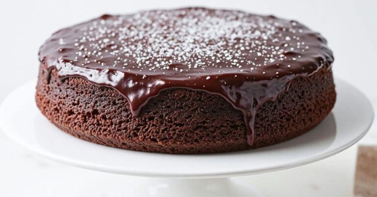 Small chocolate cake with glossy ganache topping and powdered sugar on a white plate