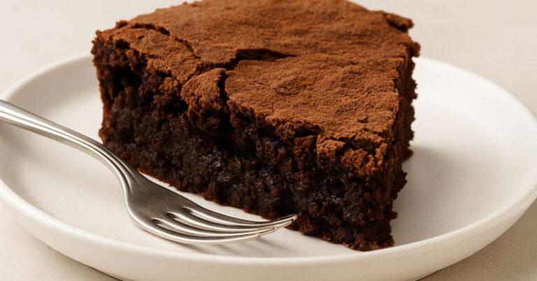 A single slice of rich, fudgy brownie cake on a white plate, with a moist interior and cocoa-dusted cracked top, styled on a beige background.
