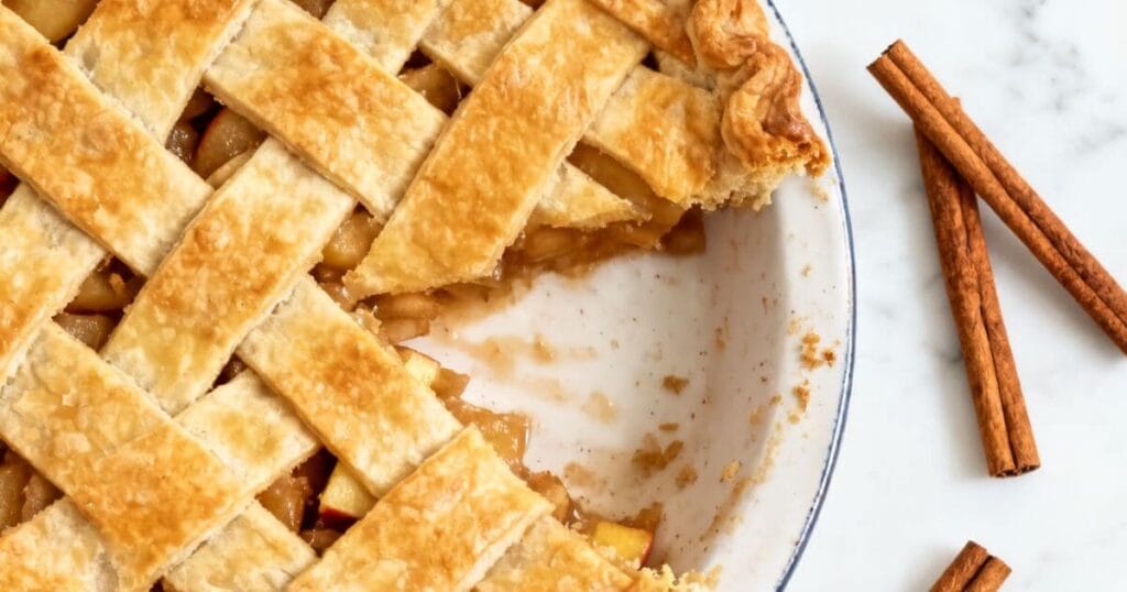 Overhead view of the best apple pie recipe featuring a golden lattice crust and 1 secret ingredient, with a slice removed to show the spiced apple filling and cinnamon sticks on the side