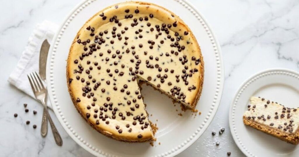 A baked chocolate chip cheesecake with a slice cut out, sitting on a white beaded cake stand next to a separate plate with a slice of cheesecake, a fork, a knife, and scattered mini chocolate chips.