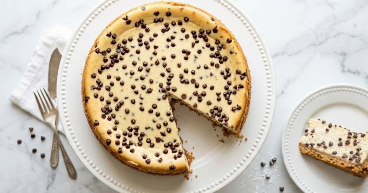 A baked chocolate chip cheesecake with a slice cut out, sitting on a white beaded cake stand next to a separate plate with a slice of cheesecake, a fork, a knife, and scattered mini chocolate chips.
